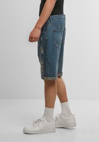 Shorts en denim bleu moyen avec des patchs déchirés et des ourlets roulés, associés à des baskets blanches et des chaussettes blanches de longueur mi-mollet.
