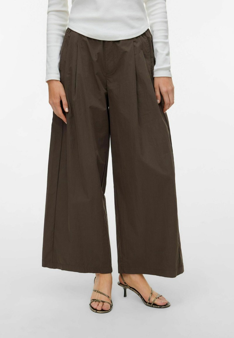 Femme portant un pantalon pattes d'éléphant marron, un haut blanc à manches longues et des sandales à talons avec lanières, debout sur un fond blanc uni.