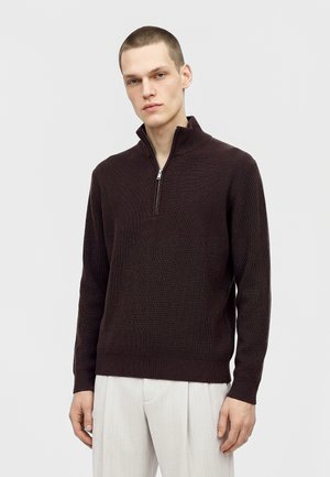 Modèle masculin portant un pull côtelé marron foncé à demi-zip et un pantalon blanc, sur un fond clair uni.