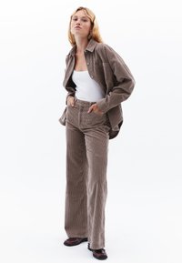 Bruine corduroy overhemd en bijpassende high-waisted broek, met wijde pijpen en een gestructureerd oppervlak. Model draagt er een witte tanktop onder.