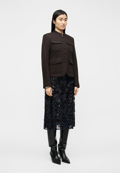 Γυναίκα που φοράει σκούρο καφέ κουμπωτό σακάκι με τσέπες, μαύρη μίντι φούστα με παγιέτες και μαύρες μυτερές μπότες μέχρι το γόνατο, σε απλό φόντο.