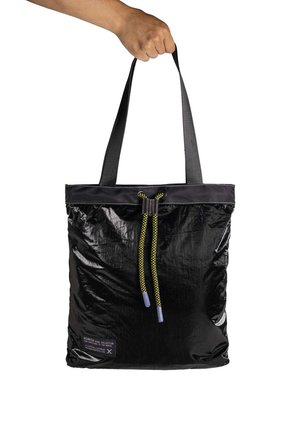 Hand hält eine schwarze, glänzende Tragetasche mit Zugband-Verschluss aus gelber und schwarzer Schnur sowie schwarzen Schulterriemen.