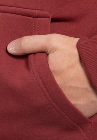 Bordový svetr s měkkou texturou, s přední kapsou a rukou položenou uvnitř, která zdůrazňuje detaily švu a tkaniny.