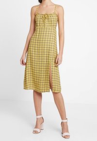 Vestido amarillento con estampado de cuadros, tirantes ajustables, un frente fruncido, lazos en el escote y una abertura lateral. Combinado con tacones de color claro.