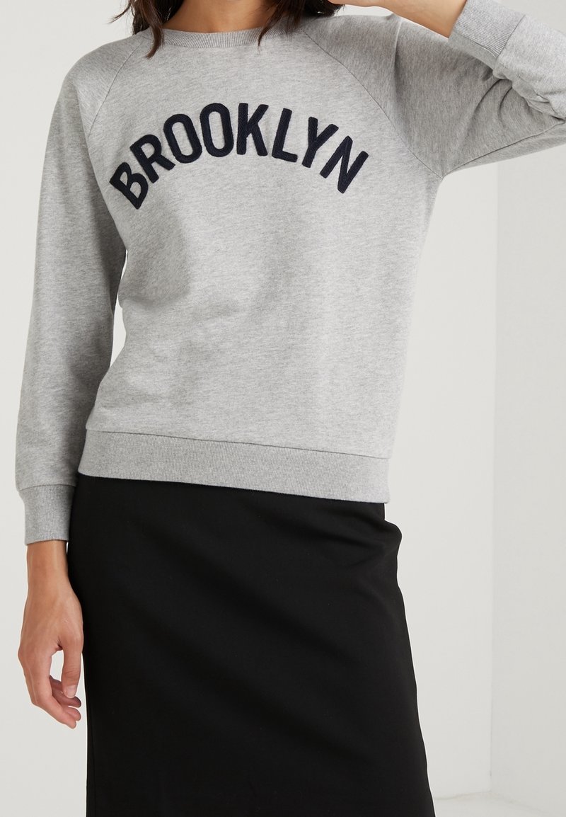 Šedá mikina s vyšitým nápisem "BROOKLYN" v námořnické modři, raglánové rukávy a žebrované manžety. Nositelná přes černou sukni, se casual vzhledem.