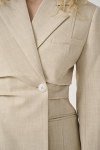 Veste beige ajustée en tissu texturé, dotée d'un design croisé, d'une fermeture à un seul bouton et d'une poche poitrine avec un accent subtile.