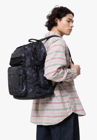 Svart ryggsäck med mönstrad textur och flera fack, bärs på ryggen. Bäraren har på sig en randig skjorta i rosa, grön och vit.