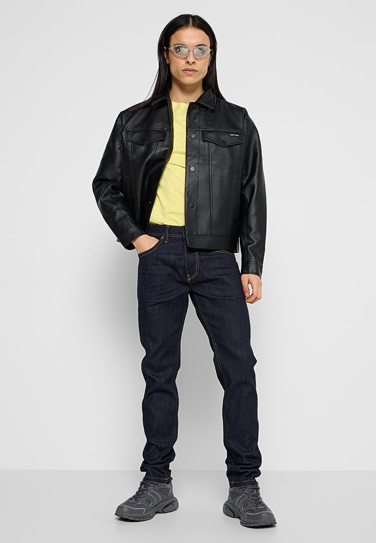 Schwarze Kunstlederjacke mit Knopfverschluss, darunter ein gelbes T-Shirt, dunkle Denim-Jeans und graue Sneakers. Eine klare Brille rundet das Outfit ab.