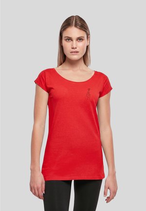 Femme aux cheveux lisses châtain clair portant un t-shirt rouge à manches courtes avec un petit geste de la main et un motif cœur sur la poitrine.