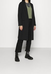 Zwarte lange jas over een groen gebreid vest en een gestreept topje, gecombineerd met zwarte broek en enkellaarsjes. Gladde stof, getailleerde pasvorm.