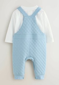 Svetlo modra prešita kombinezona z nastavljivimi naramnicami, v kombinaciji z belim dolgim rokavom spodaj. Mehka tkanina, sproščen kroj, rebrasti manšeti.