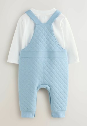 Salopette matelassée bleu clair avec bretelles réglables, portée sur une chemise blanche à manches longues. Tissu doux, coupe décontractée, poignets côtelés.