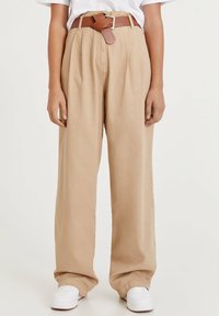 Pantalon large beige en tissu léger, avec une taille haute, des plis et une ceinture en cuir marron. Des baskets blanches complètent le look.