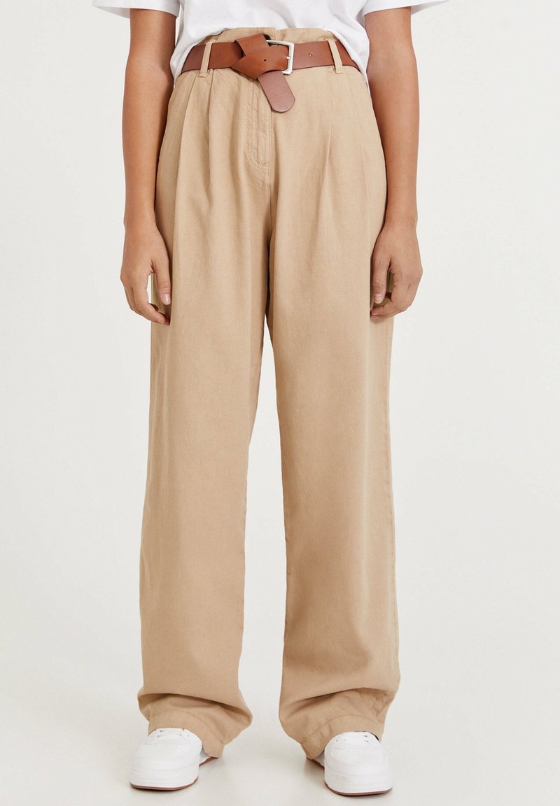 Pantalon large beige en tissu léger, avec une taille haute, des plis et une ceinture en cuir marron. Des baskets blanches complètent le look.