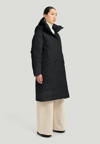 Svart ytterjacka med huva, med en minimalistisk design, medellång längd och en dold knappslutning. Ett par vida beiga byxor bärs under.