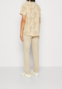 Chemise à manches courtes, beige clair, avec un motif de feuille de palmier, associée à un pantalon beige et des baskets blanches avec un logo beige.