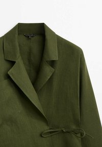 Olive green linen blazer featuring a notched collar, single pocket, and a tie detail at the waist for adjustable fit. Smooth texture.