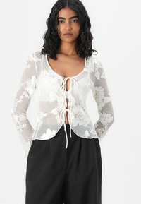Sheer white blouse with floral embroidery, round neck, tie-front detail, and long sleeves; worn over black pants. Delicate texture and design.