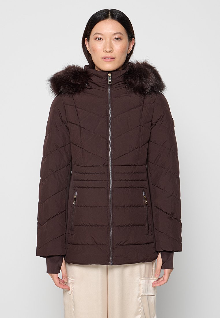 Vrouw in een donkerbruine, gewatteerde winterjas met een met bont afgewerkte capuchon, vooraan een rits en met ritszakken, staande tegen een lichte achtergrond.