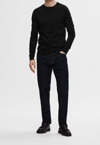 Pull noir à col rond, associé à un jean bleu foncé. Le mannequin porte des chaussures noires, debout devant un fond neutre.