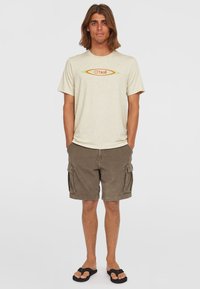 Beige Baumwoll-T-Shirt mit einem orangefarbenen und grünen O'Neill-Logo; kombiniert mit braunen Cargo-Shorts mit Seitentaschen und schwarzen Flip-Flops.