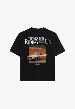 Jonge man die een zwart T-shirt met witte tekst draagt, een donkergroene bandana om zijn nek geknoopt, een zwarte pet met een oranje logo en groene shorts.