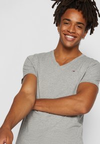 Camiseta gris de cuello en V en tela suave, con mangas cortas. Cuenta con un pequeño logo en el pecho. Textura suave, ajuste casual.