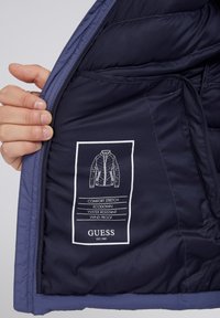 Intérieur d'une doudoune navy montrant une texture lisse, une étiquette imprimée avec des instructions d'entretien, et des caractéristiques : confort extensible, résistant à l'eau, coupe-vent.