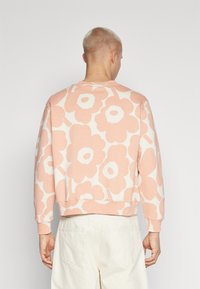 Felpa rosa chiaro con grandi motivi floreali bianchi, maniche lunghe e orlo a costine. Il tessuto sembra morbido e confortevole.