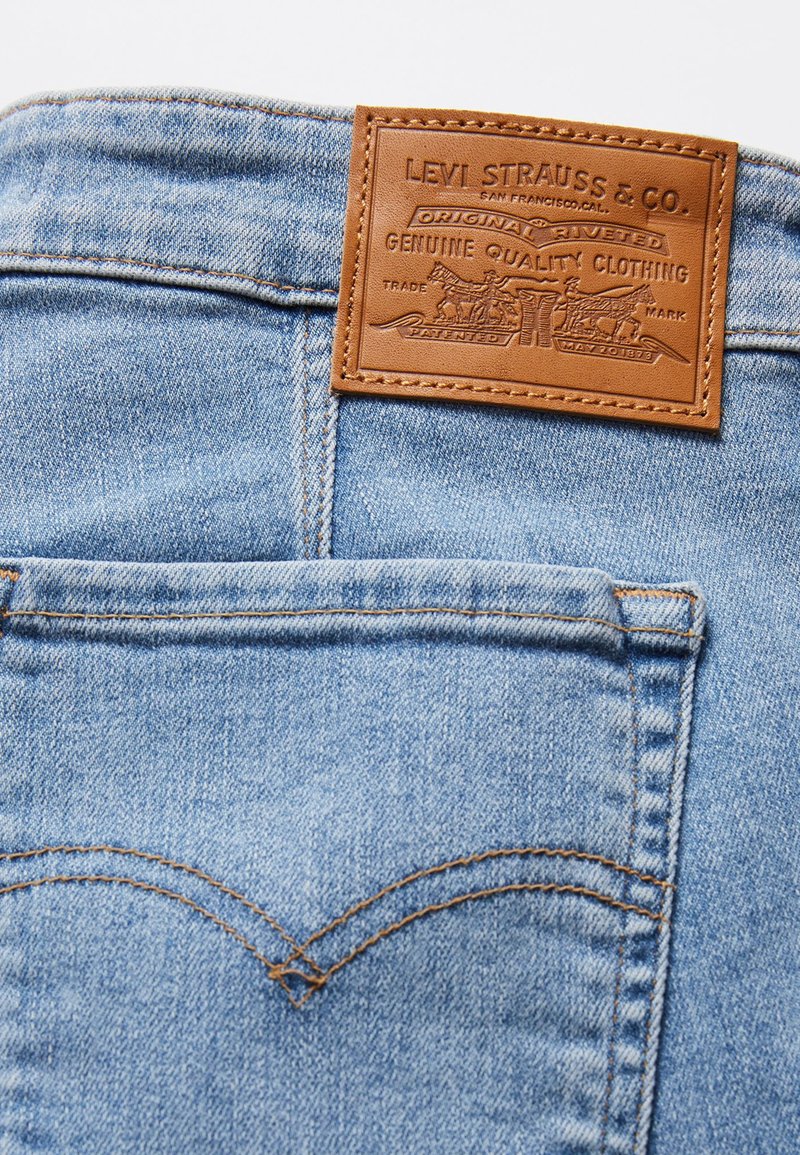 Los jeans de mezclilla azul claro presentan un parche de cuero marrón con texto en relieve y un diseño de herradura. La costura es visible en el bolsillo trasero.