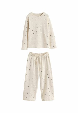 Junges Mädchen im cremefarbenen Pyjama mit kleinen Punkten steht barfuß und lehnt an einem grünen modernen Stuhl in einem sanft beleuchteten Raum mit beigem Teppich.