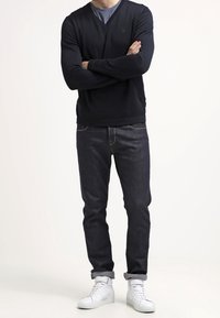 Muž stojí s překříženýma rukama, oblečený v černém svetr s výstřihem do V, modré košili, tmavých džínách a bílých teniskách na světlém pozadí.