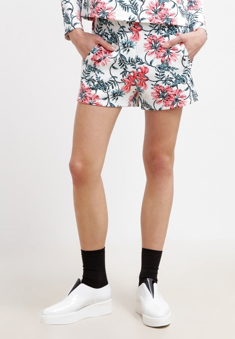 Personne portant un short blanc et un haut assorti avec un imprimé floral rose, des chaussettes noires et des chaussures blanches à plateforme sans lacets. Mains dans les poches.