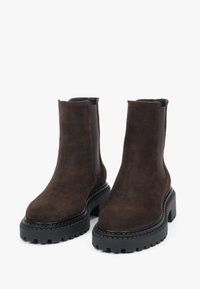 Brązowe botki ze skóry zamszowej z czarną gumową platformą, elastycznymi panelami po bokach i zaokrąglonym noskiem. Dekoracyjne przeszycia wzdłuż podeszwy.