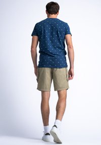 Tricou bleumarin cu un model de frunze verzi, asociat cu pantaloni scurți kaki. Modelul poartă adidași albi și șosete scurte, întorcându-se cu fața de la privitor.