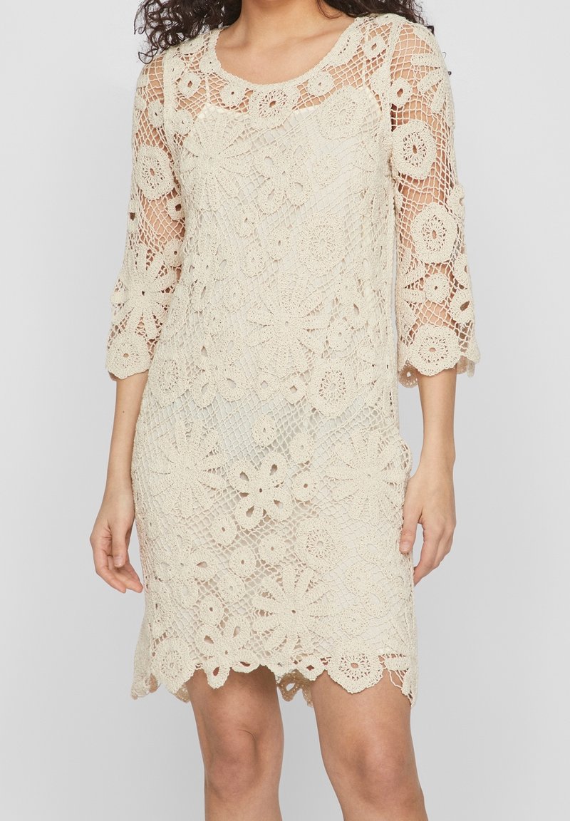 Femme portant une robe beige en crochet longueur genou avec motifs floraux et manches trois-quarts, debout devant un fond uni.