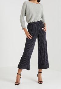 Dunkel gestreifte Hose mit elastischem Bund, weitem Bein und umgeschlagenem Saum. Getragen mit einem hellgrauen Pullover und schwarzen Riemchen-Heels.
