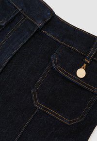 Ciemne dżinsy z złotym przeszyciem i pojedynczą tylną kieszenią z okrągłym złotym guzikiem. Tkanina ma fakturę z subtelnym połyskiem.