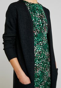 Cardigan gris foncé à coupe ample avec des poignets côtelés, porté sur une robe verte ornée d'un motif à pois coloré en rose, blanc et noir.