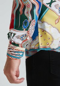 Gros plan sur une manche de chemise à motifs colorés avec un poignet boutonné portée avec un pantalon noir devant un fond uni.