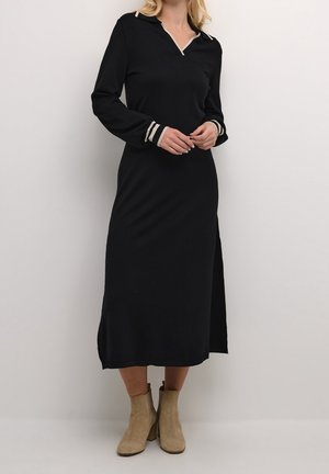 Femme portant une longue robe noire avec un col et des poignets rayés, associée à des bottines beiges, debout sur un fond blanc.
