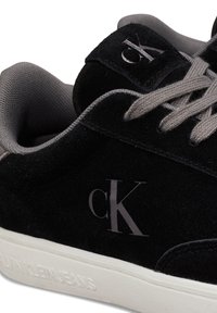 Schwarze Wildleder-Sneaker mit grauen Schnürsenkeln, versehen mit einem dezenten Logo auf der Zunge und einer weißen Gummisohle, die mit "CALVIN KLEIN JEANS" geprägt ist.