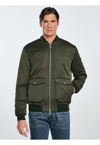 Olivgrüne Bomberjacke mit durchgehendem Reißverschluss, gerippten Bündchen, zwei Fronttaschen und Logodetail auf der Brust. Glattes Material mit leichtem Glanz.