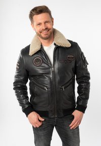 Schwarze Leder-Bomberjacke mit einem Kragen aus Kunstfaser-Schaffell, zwei Reißverschluss-Fronttaschen und gestickten Patches auf Brust und Ärmeln.