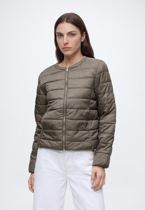 Kvinde iført en khaki quiltet jakke med lynlås og hvide bukser, stående mod en ensfarvet lys baggrund med et neutralt udtryk.