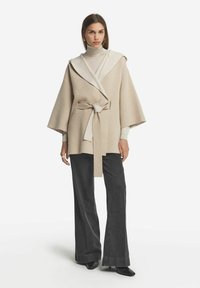 Beige inneslutningsjacka med huva, strukturerad tyg, vida ärmar och bälte, lager på en ljus polotröja och i kombination med mörka byxor.