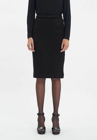 Jupe crayon noire avec une taille texturée, en tissu lisse. Associée à des chaussures à talons hauts noires et des collants transparents.