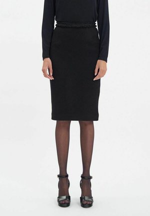 Jupe crayon noire avec une taille texturée, en tissu lisse. Associée à des chaussures à talons hauts noires et des collants transparents.