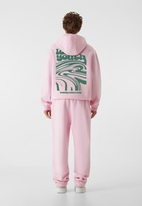 Un sweat à capuche rose avec un graphique vert dans le dos, associé à un pantalon de survêtement rose assorti et des baskets blanches. Matière douce et coupe décontractée.