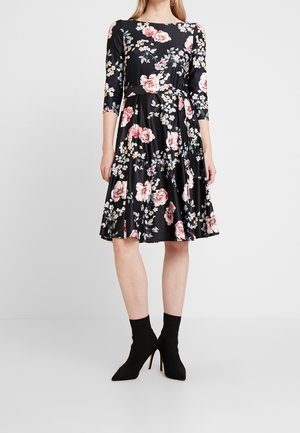 Kvinde iført en sort kjole til knæet med pink og hvidt blomsterprint og trekvartlange ærmer, kombineret med sorte ankelstøvler med høje hæle.